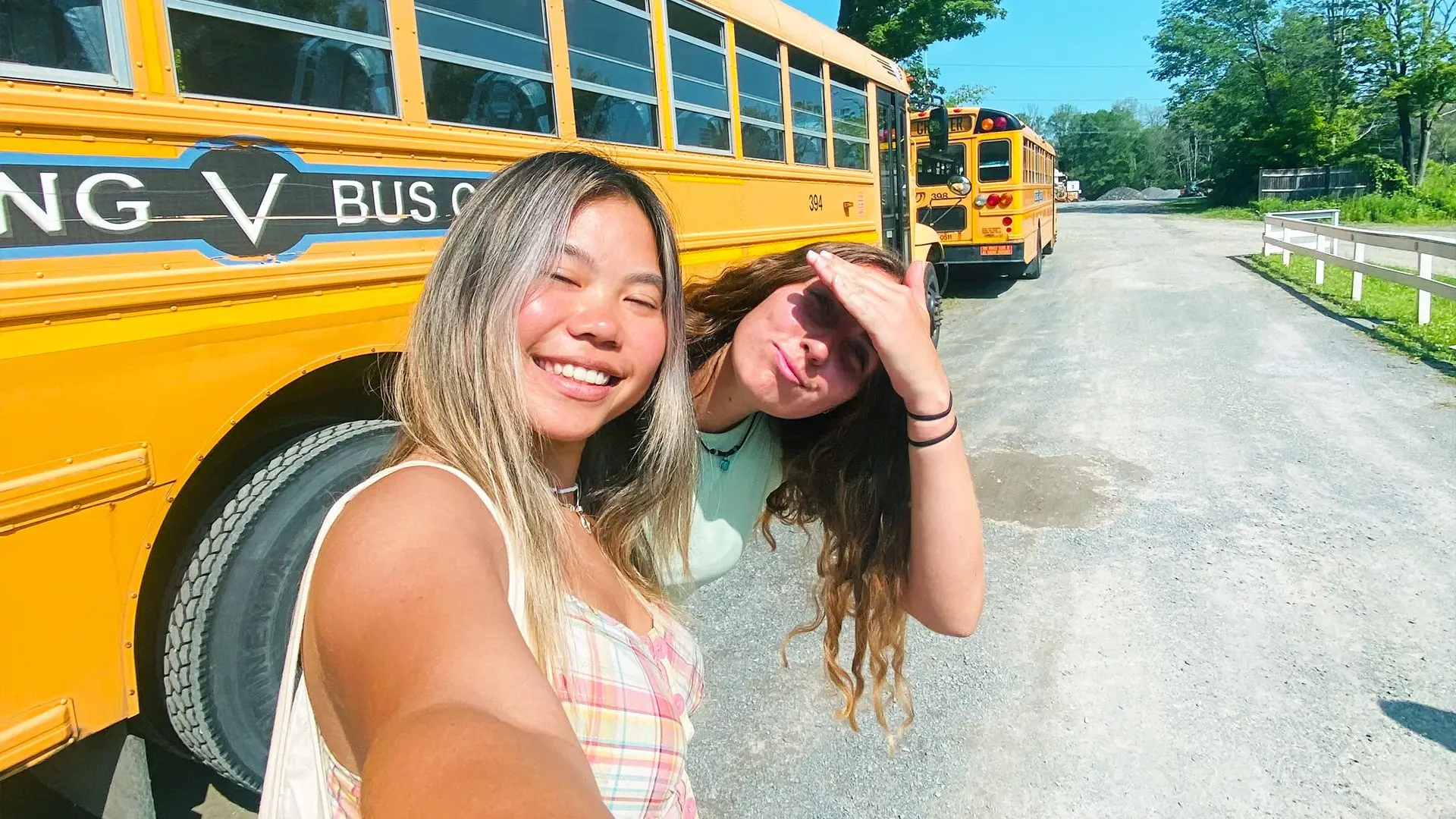 Two girls taking a selfie in front of an American yellow school bus at summer camp.