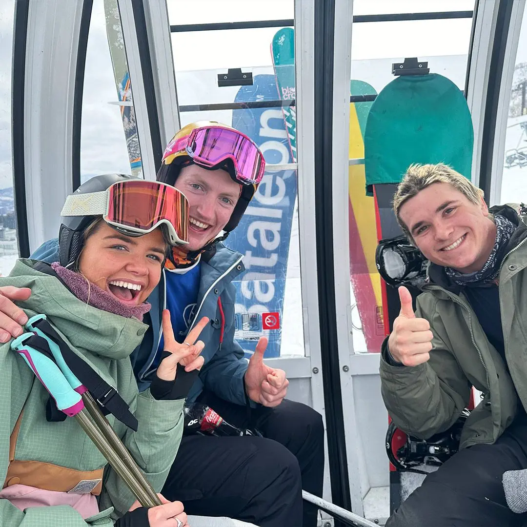 Three friends in a ski lift, two male and one female. They're all wearing ski gear and have bright ski goggles on. They're all smiling and posing for the camera with thumbs up and peace signs.