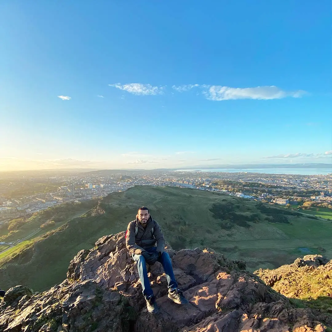 Guy sits on mountain beside Edinburgh on sunny morning.